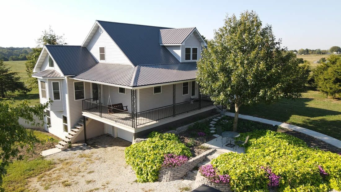 Aerial view of a white farmhouse with dark standing seam metal roof, dormers and raised deck