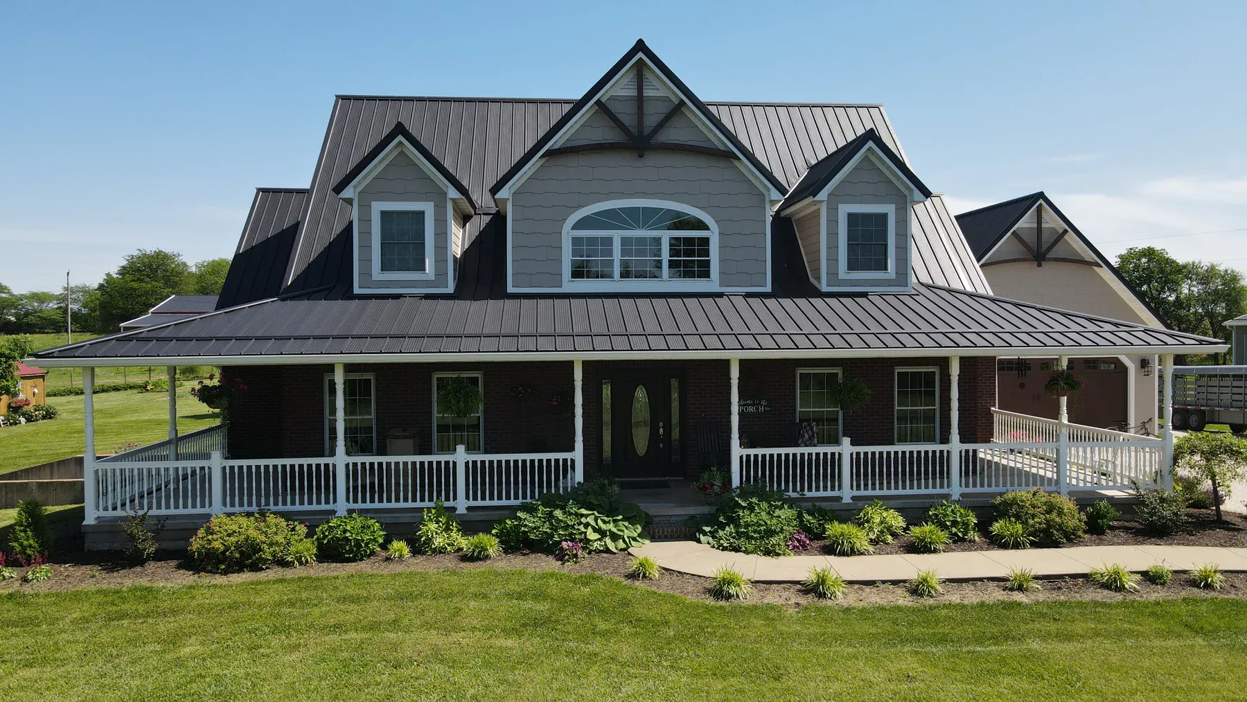 Grey two-story home with brown standing seam metal roof, triple dormers and brick wrap-around porch