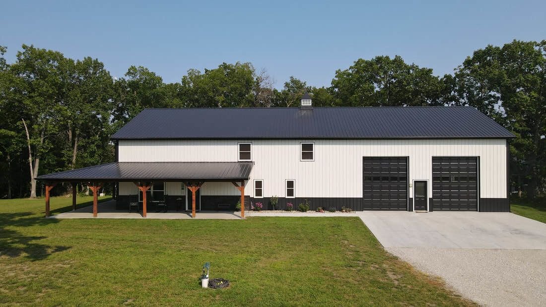 White steel building with black metal roof, black wainscot, timber-post porch and two tall garage doors