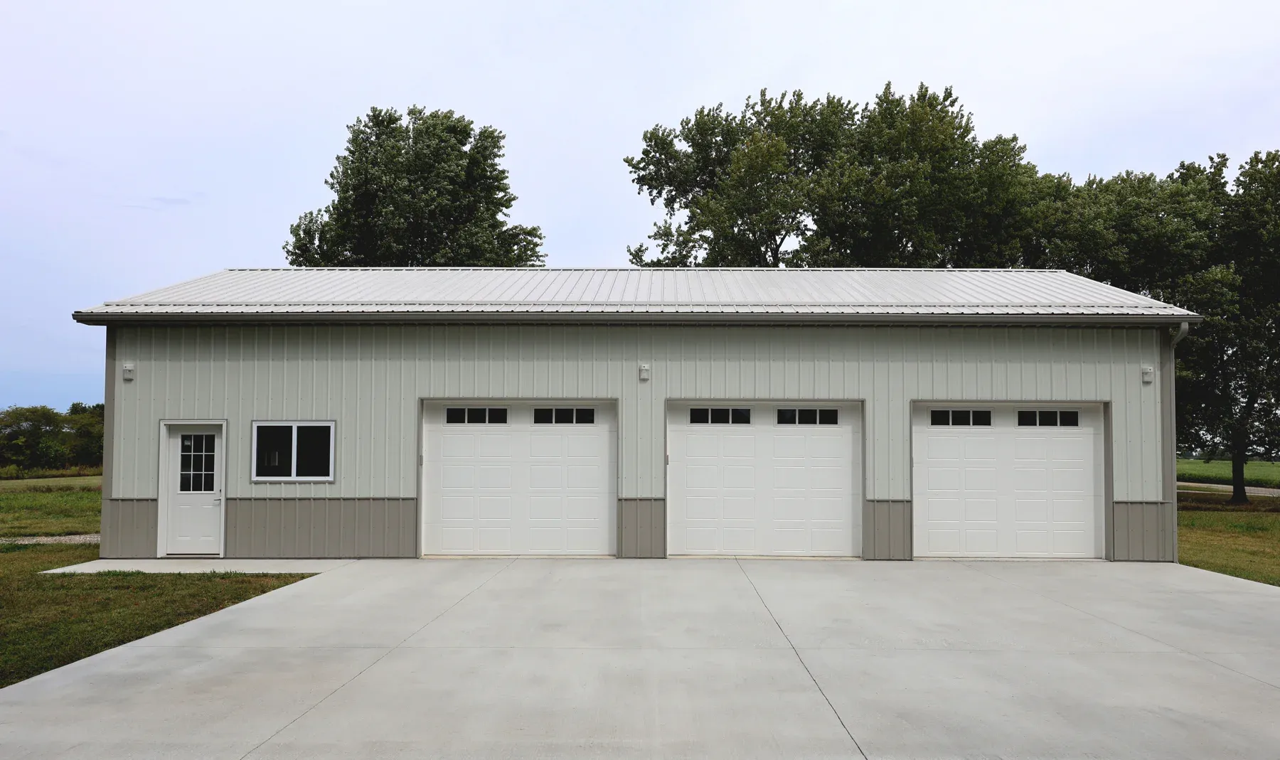 Light grey steel garage with tan wainscot, silver metal roof and three white overhead doors