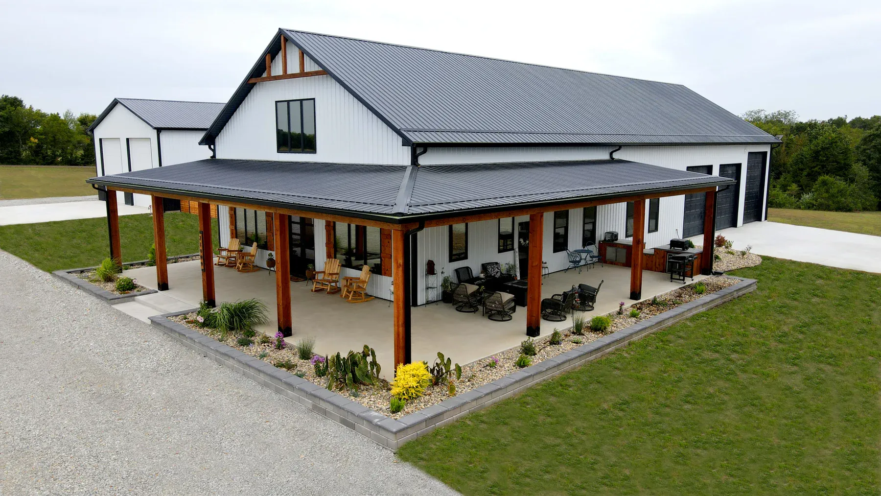 White steel barndominium with black metal roof and wide timber-post wrap-around porch