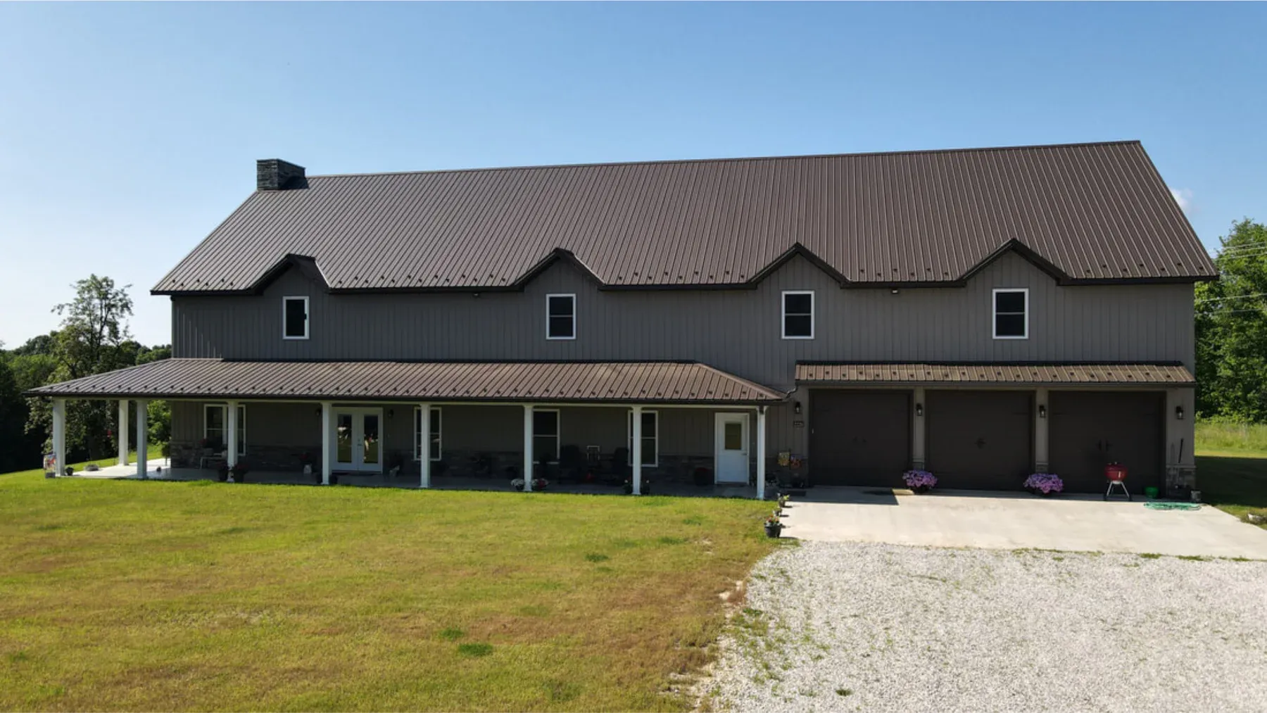 Two-story brown steel home with dormers, wrap-around porch and three-bay garage